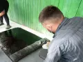 Zwei Personen arbeiten daran, den Boden abzudichten. Eine Frau gießt schwarze Flüssigkeit aus einem Eimer, während ein Mann eine Rolle Bitumenbahn ausrollt.