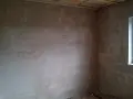 Unverputzte Innenwand mit Fensterecke und sichtbaren Deckenbalken.