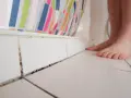 Schwarze Flecken in der Dusche, wo die Fliesen auf den Boden treffen. Ein Fuß steht daneben, die bunte Duschvorhang ist im Hintergrund.