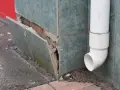 Beschädigte Sockelabdichtung am Haus. Fliesen lösen sich, darunter Ziegelmauerwerk. Ein Fallrohr leitet Wasser ab.