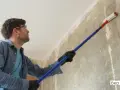 Mann mit Schutzbrille trägt mit langem Stiel und Rolle Öko Tiefengrund auf eine rohe Wand auf.