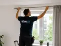 Mann bringt eine Gardinenstange an. Er steht auf einer Leiter und richtet die Stange über dem Fenster aus.