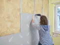 Frau schraubt Rigipsplatten an eine Wand mit Dämmung. Die Platten werden auf Metallschienen befestigt.