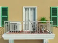 Balkon eines Mehrfamilienhauses mit Klimaanlage, Tisch und Stuhl. Grüne Fensterläden und eine Tür mit Vorhängen.