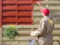 Person in beige Overall lackiert Holzzaun rot. Das geht auch ohne Schleifen, wie die Marke "Moose Färg" zeigt.