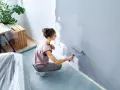 Frau malt Wand grau mit Farbroller. Sie kniet auf dem Boden, der mit Folie geschützt ist. So kann man richtig streichen.