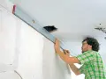 Mann richtet eine Wasserwaage an der Decke aus, um die Wand mit Rigips zu verkleiden. Kabel hängen aus einer Deckenöffnung.