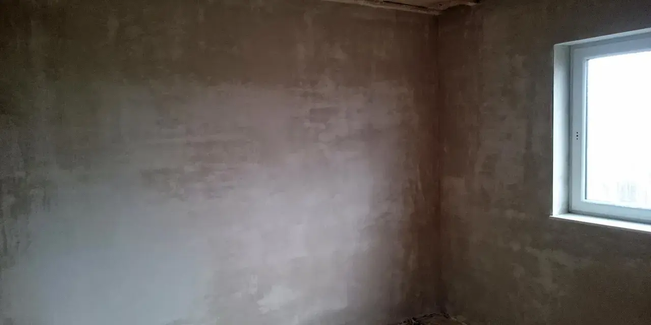 Unverputzte Innenwand mit Fensterecke und sichtbaren Deckenbalken.