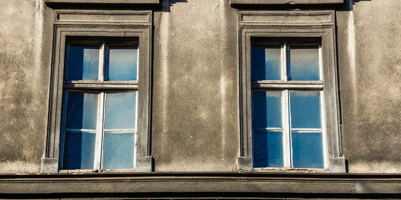 Zwei alte Fenster mit blauen Scheiben an einer verwitterten Fassade. Eine energetische Sanierung der Fenster würde sich hier lohnen.