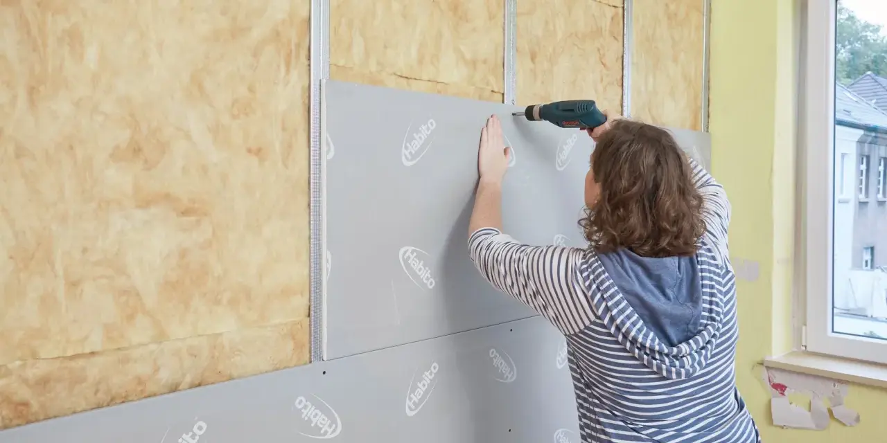 Frau schraubt Rigipsplatten an eine Wand mit Dämmung. Die Platten werden auf Metallschienen befestigt.
