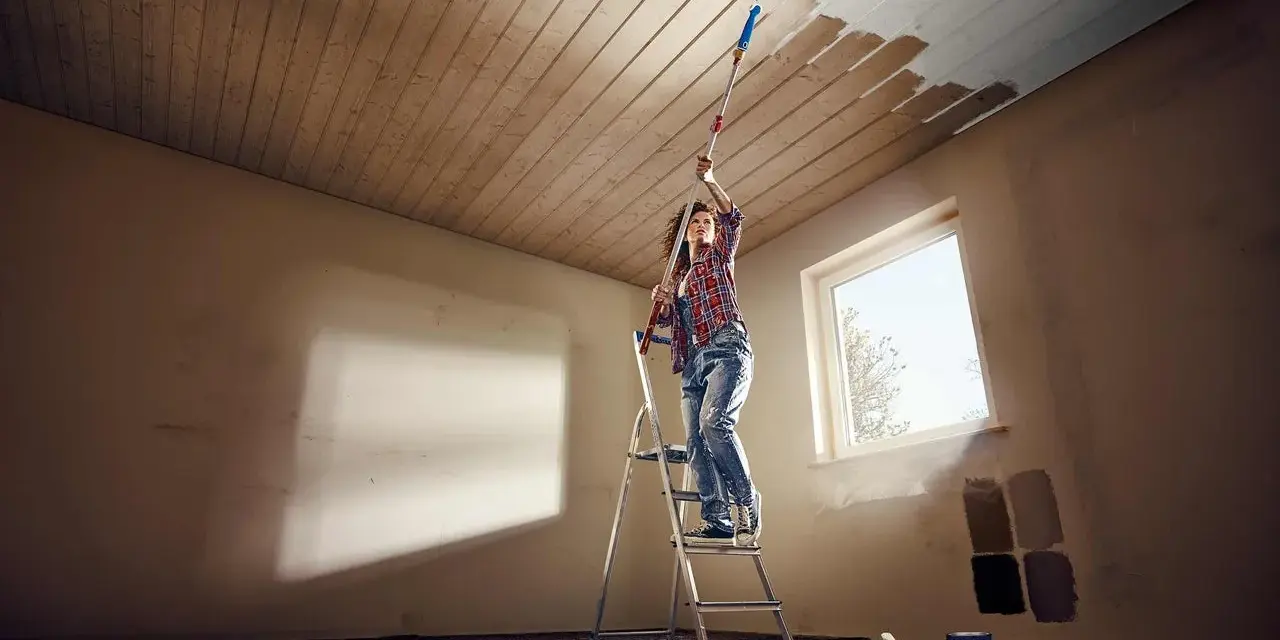Frau streicht mit einer Teleskopstange und Rolle die Holzpaneele an der Decke weiß.
