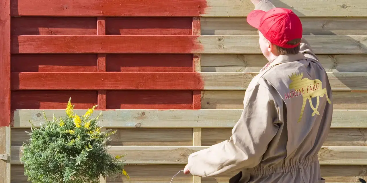 Person in beige Overall lackiert Holzzaun rot. Das geht auch ohne Schleifen, wie die Marke "Moose Färg" zeigt.