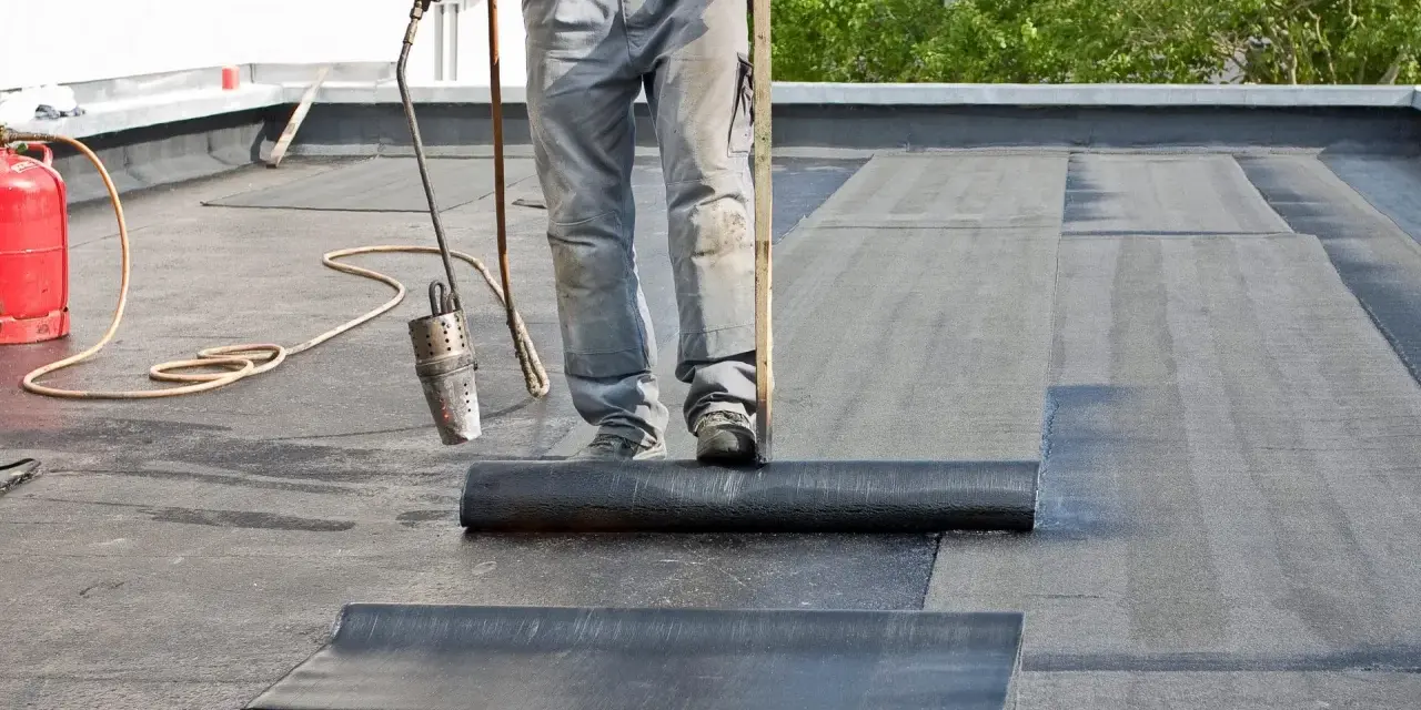 Arbeiter verlegt mit Brenner und Rolle Dachpappe. Die Hitze schmilzt die Unterseite, um eine wasserdichte Abdichtung zu schaffen.