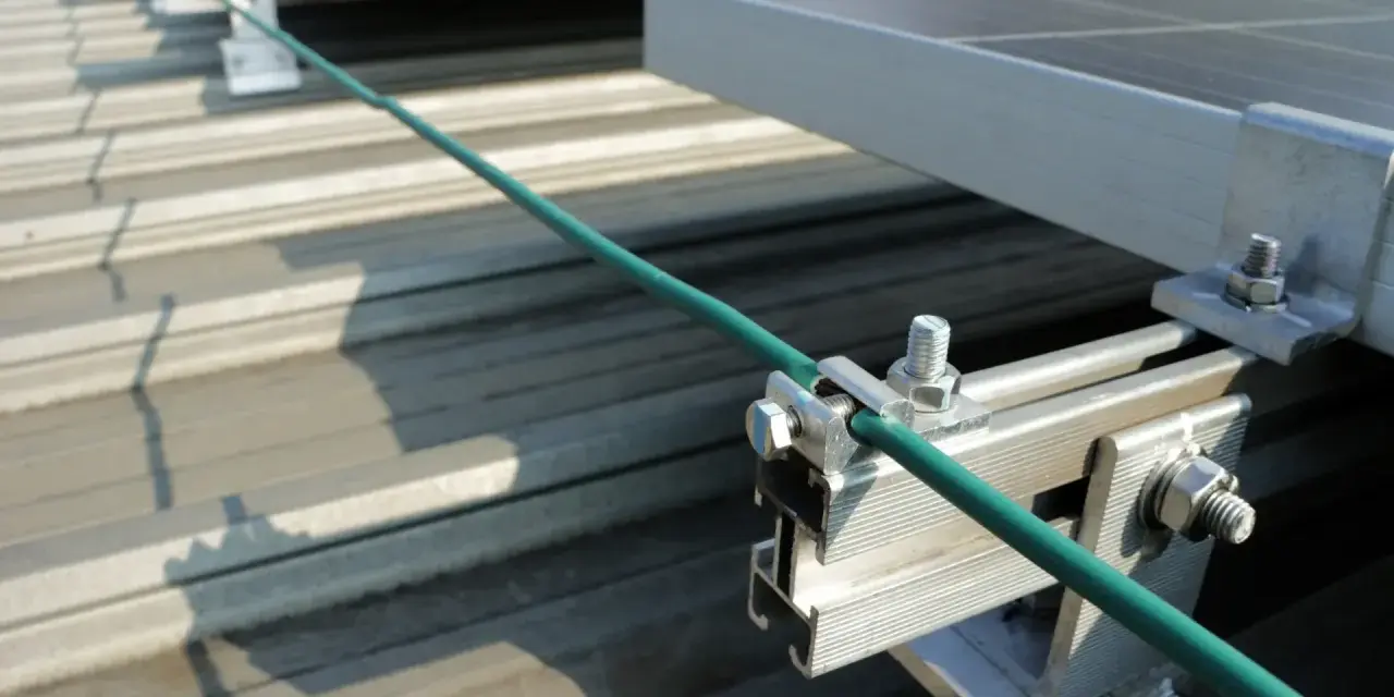 Detailaufnahme der Erdung einer PV-Anlage. Ein grünes Kabel wird sicher mit einer Klemme an der Aluminiumkonstruktion befestigt, die die Solarmodule auf dem Dach hält.