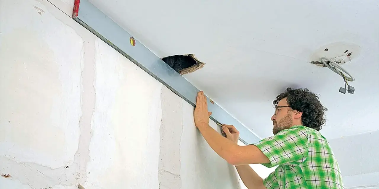 Mann richtet eine Wasserwaage an der Decke aus, um die Wand mit Rigips zu verkleiden. Kabel hängen aus einer Deckenöffnung.