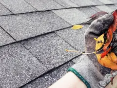 Gefüllte Regenrinne mit Laub und Schmutz, die eine Verstopfung verursacht. Eine Hand in einem Handschuh entfernt das Material.