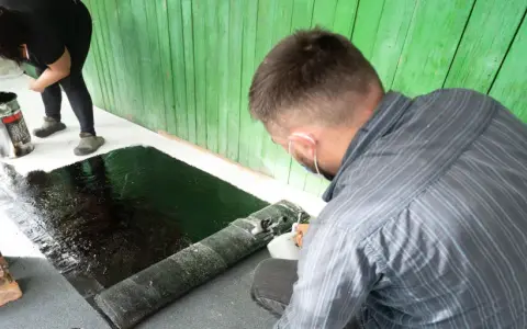 Zwei Personen arbeiten daran, den Boden abzudichten. Eine Frau gießt schwarze Flüssigkeit aus einem Eimer, während ein Mann eine Rolle Bitumenbahn ausrollt.