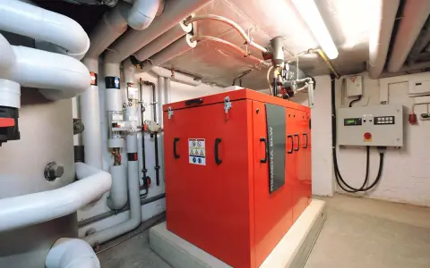 Rotes Blockheizkraftwerk im Keller eines Mehrfamilienhauses, umgeben von Rohren und einer Steuereinheit.