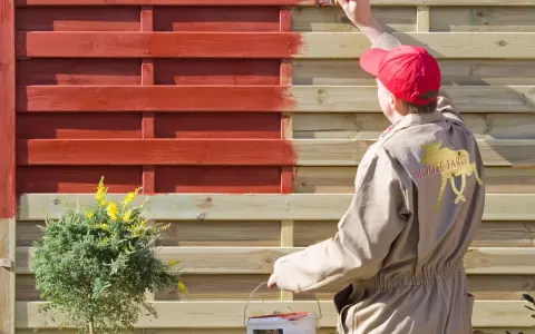 Person in beige Overall lackiert Holzzaun rot. Das geht auch ohne Schleifen, wie die Marke "Moose Färg" zeigt.
