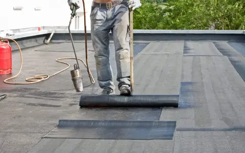 Arbeiter verlegt mit Brenner und Rolle Dachpappe. Die Hitze schmilzt die Unterseite, um eine wasserdichte Abdichtung zu schaffen.