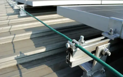 Detailaufnahme der Erdung einer PV-Anlage. Ein grünes Kabel wird sicher mit einer Klemme an der Aluminiumkonstruktion befestigt, die die Solarmodule auf dem Dach hält.