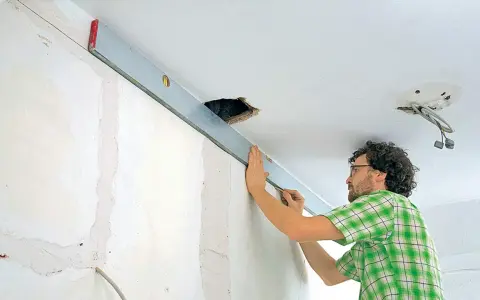 Mann richtet eine Wasserwaage an der Decke aus, um die Wand mit Rigips zu verkleiden. Kabel hängen aus einer Deckenöffnung.