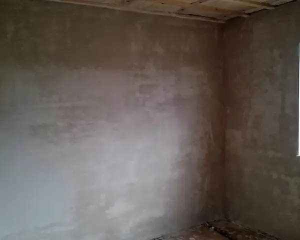 Unverputzte Innenwand mit Fensterecke und sichtbaren Deckenbalken.