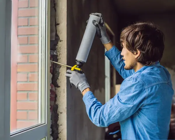 Mann mit Handschuhen und Schaumpistole dichtet den Rahmen für den Fenstertausch ab.