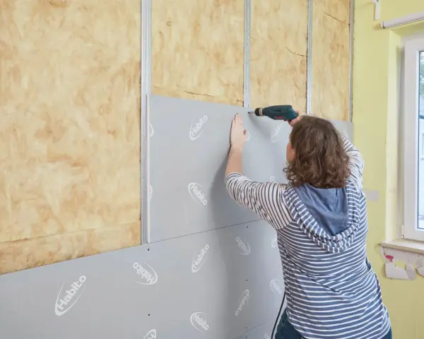 Frau schraubt Rigipsplatten an eine Wand mit Dämmung. Die Platten werden auf Metallschienen befestigt.