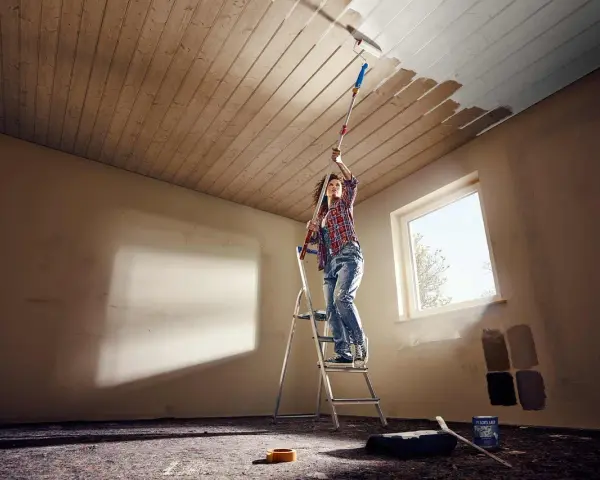 Frau streicht mit einer Teleskopstange und Rolle die Holzpaneele an der Decke weiß.