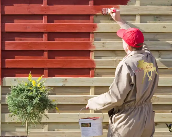 Person in beige Overall lackiert Holzzaun rot. Das geht auch ohne Schleifen, wie die Marke "Moose Färg" zeigt.
