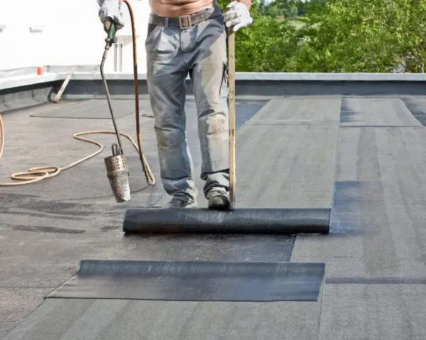 Arbeiter verlegt mit Brenner und Rolle Dachpappe. Die Hitze schmilzt die Unterseite, um eine wasserdichte Abdichtung zu schaffen.