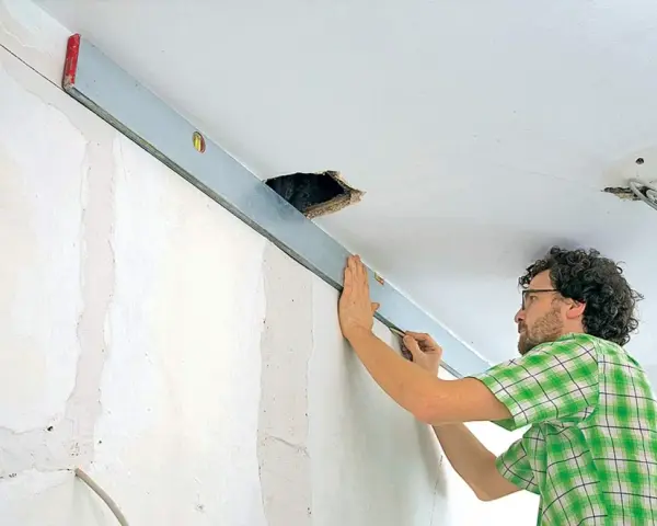 Mann richtet eine Wasserwaage an der Decke aus, um die Wand mit Rigips zu verkleiden. Kabel hängen aus einer Deckenöffnung.