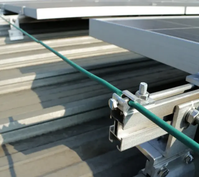 Detailaufnahme der Erdung einer PV-Anlage. Ein grünes Kabel wird sicher mit einer Klemme an der Aluminiumkonstruktion befestigt, die die Solarmodule auf dem Dach hält.