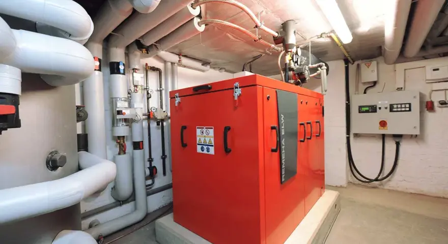 Rotes Blockheizkraftwerk im Keller eines Mehrfamilienhauses, umgeben von Rohren und einer Steuereinheit.