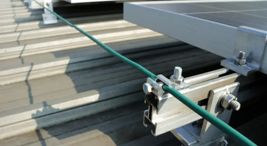Detailaufnahme der Erdung einer PV-Anlage. Ein grünes Kabel wird sicher mit einer Klemme an der Aluminiumkonstruktion befestigt, die die Solarmodule auf dem Dach hält.