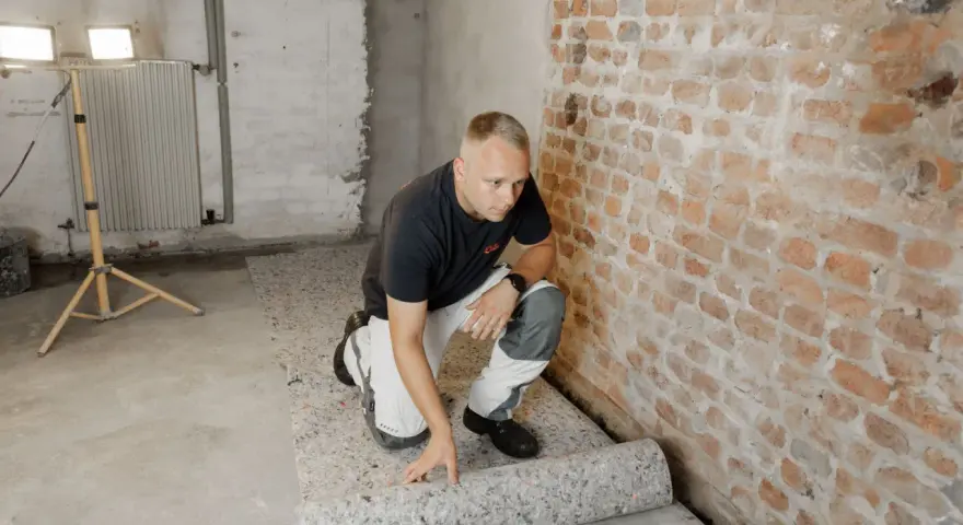 Mann rollt atmungsaktiven Bodenbelag im Keller aus. Die Ziegelwand und der Betonboden sind sichtbar.