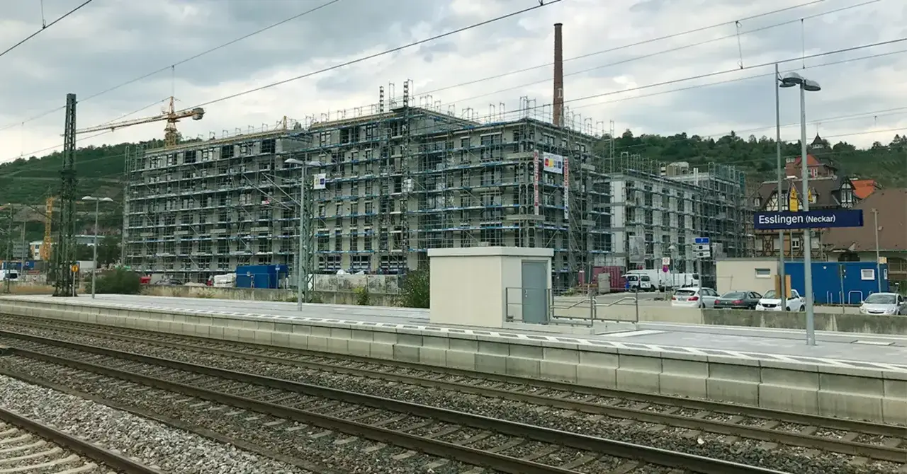 Baustelle eines Mehrfamilienhauses in Esslingen. Ein Kran ragt über das Gerüst. Im Hintergrund ein altes Backsteingebäude mit Schornstein, das vielleicht bald ein Blockheizkraftwerk beherbergen wird.