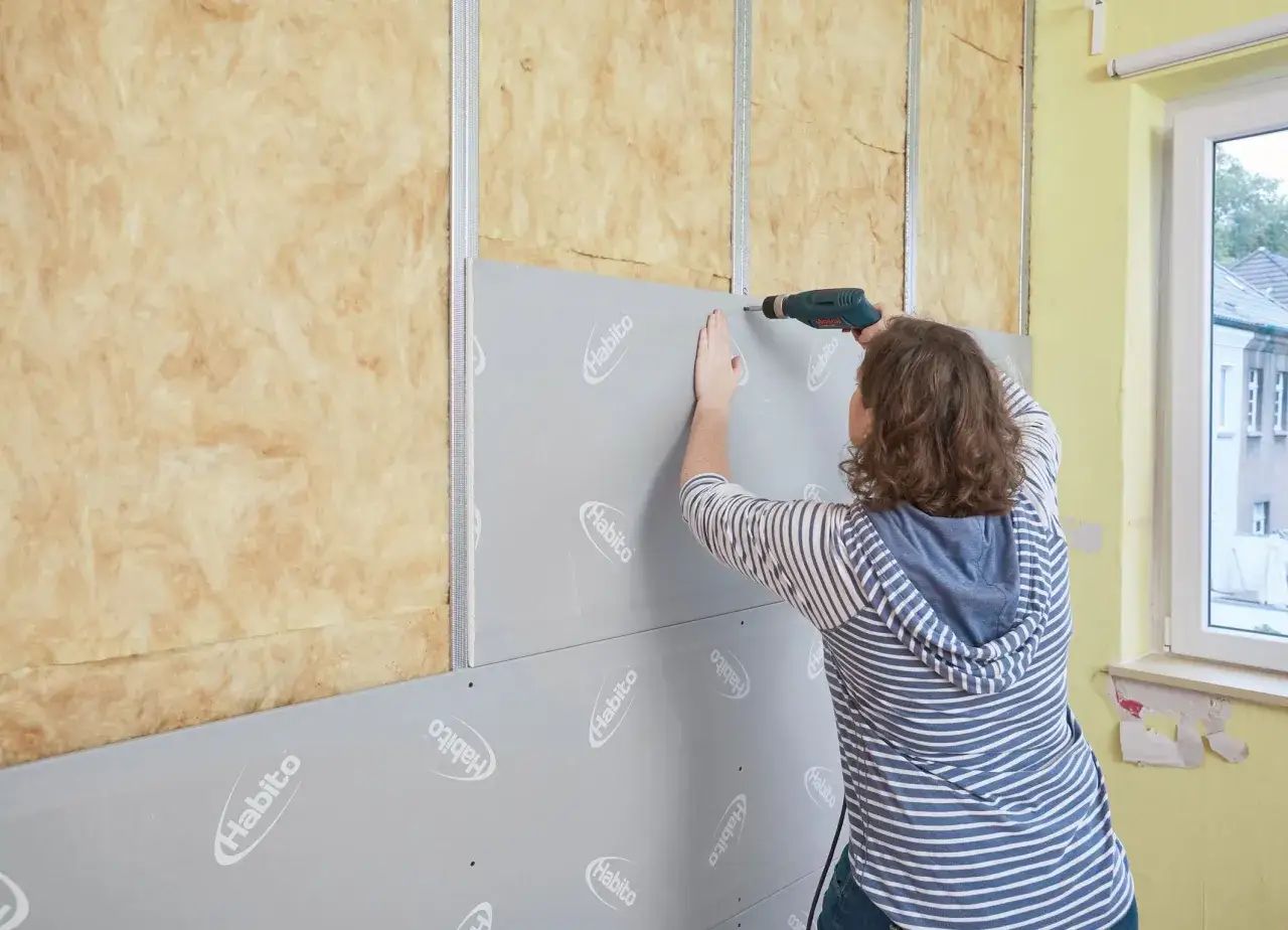 Frau schraubt Rigipsplatten an eine Wand mit Dämmung. Die Platten werden auf Metallständer montiert.