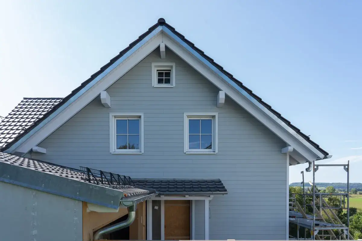 Hausfassade mit hellgrauen Eternitplatten, die auf einen neuen Anstrich warten. Gerüst und Dacharbeiten sind im Gange.