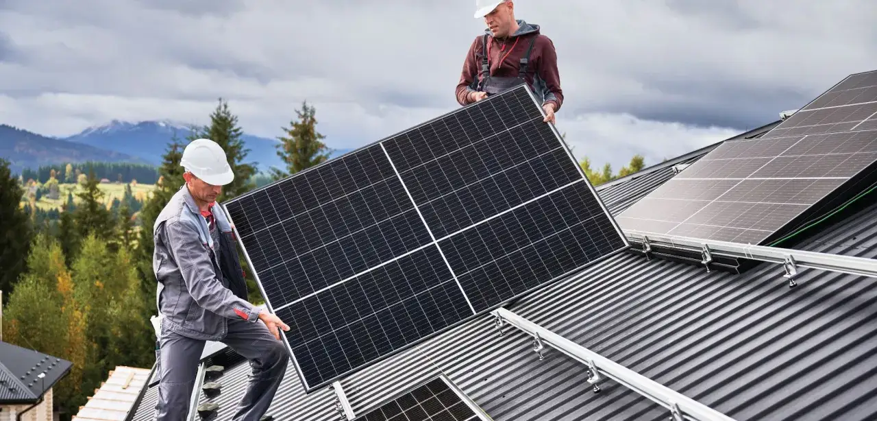 Zwei Handwerker installieren Solarmodule auf einem Dach, um die PV-Anlage zu erweitern. Im Hintergrund Berge und Bäume.
