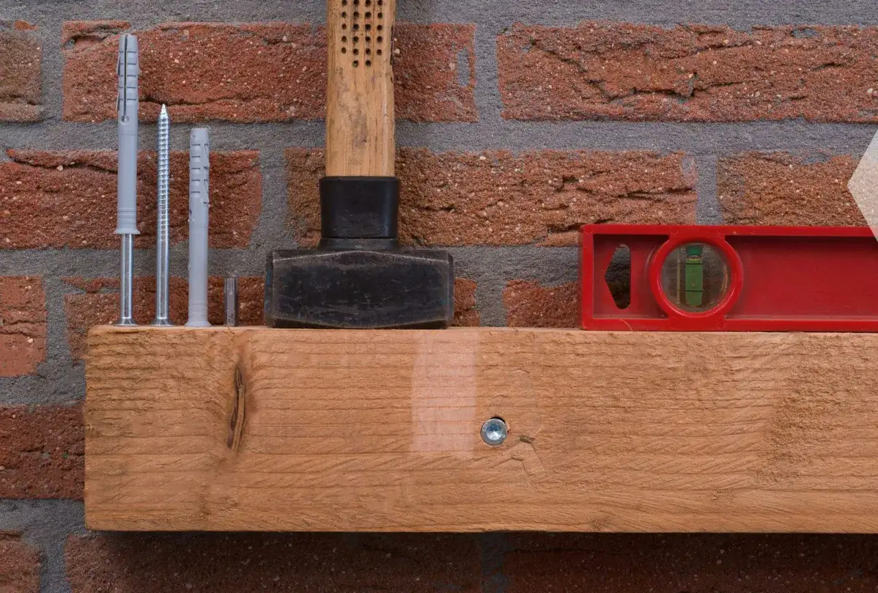 Werkzeugset zum Balken an Wand befestigen: Dübel, Schrauben, Hammer, Wasserwaage und Holzbalken vor Ziegelwand.