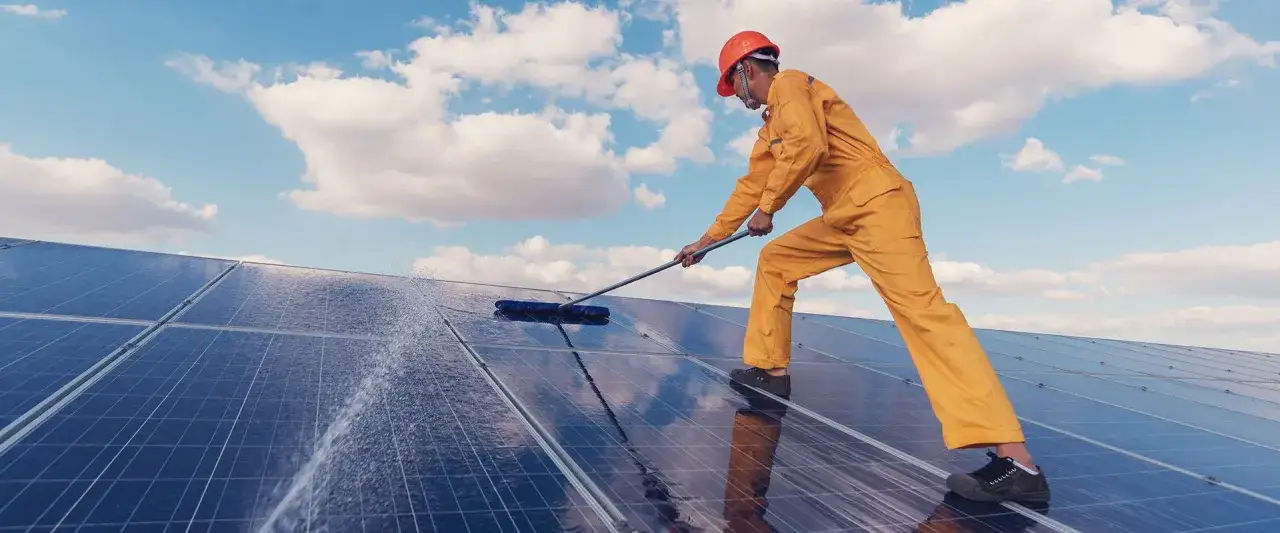 Mitarbeiter in orangefarbener Arbeitskleidung reinigt Solarmodule mit einem Besen. Die Wartung der Solaranlage ist wichtig für die Energieerzeugung.