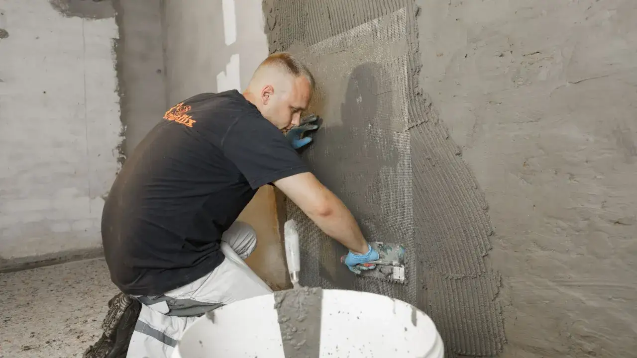 Mann dichtet eine Kellerwand von innen ab, trägt Mörtel mit einer Kelle auf ein Armierungsgewebe auf.