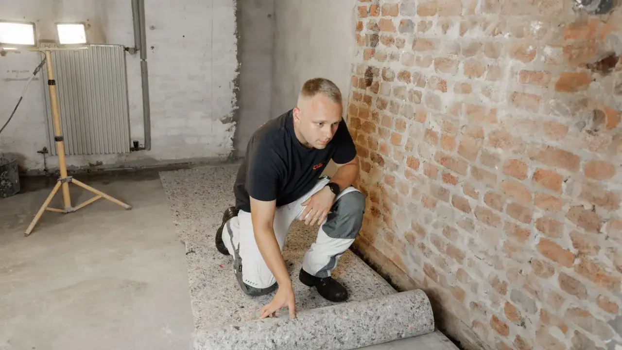 Mann rollt atmungsaktiven Bodenbelag im Keller aus. Die Ziegelwand und der Betonboden sind sichtbar.