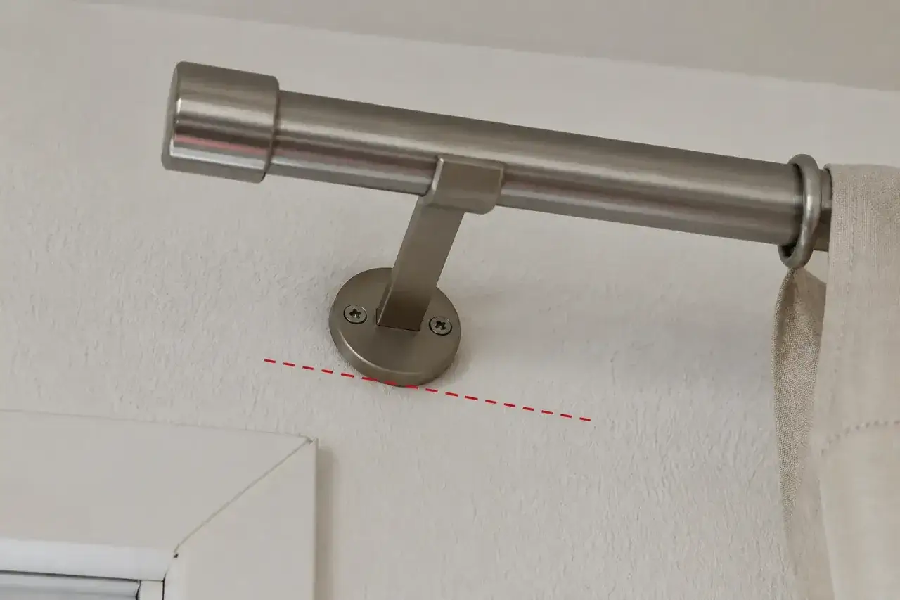 Eine Gardinenstange anbringen: Die Halterung ist mit zwei Schrauben an der Wand befestigt. Die gestrichelte rote Linie deutet auf eine leichte Neigung hin.