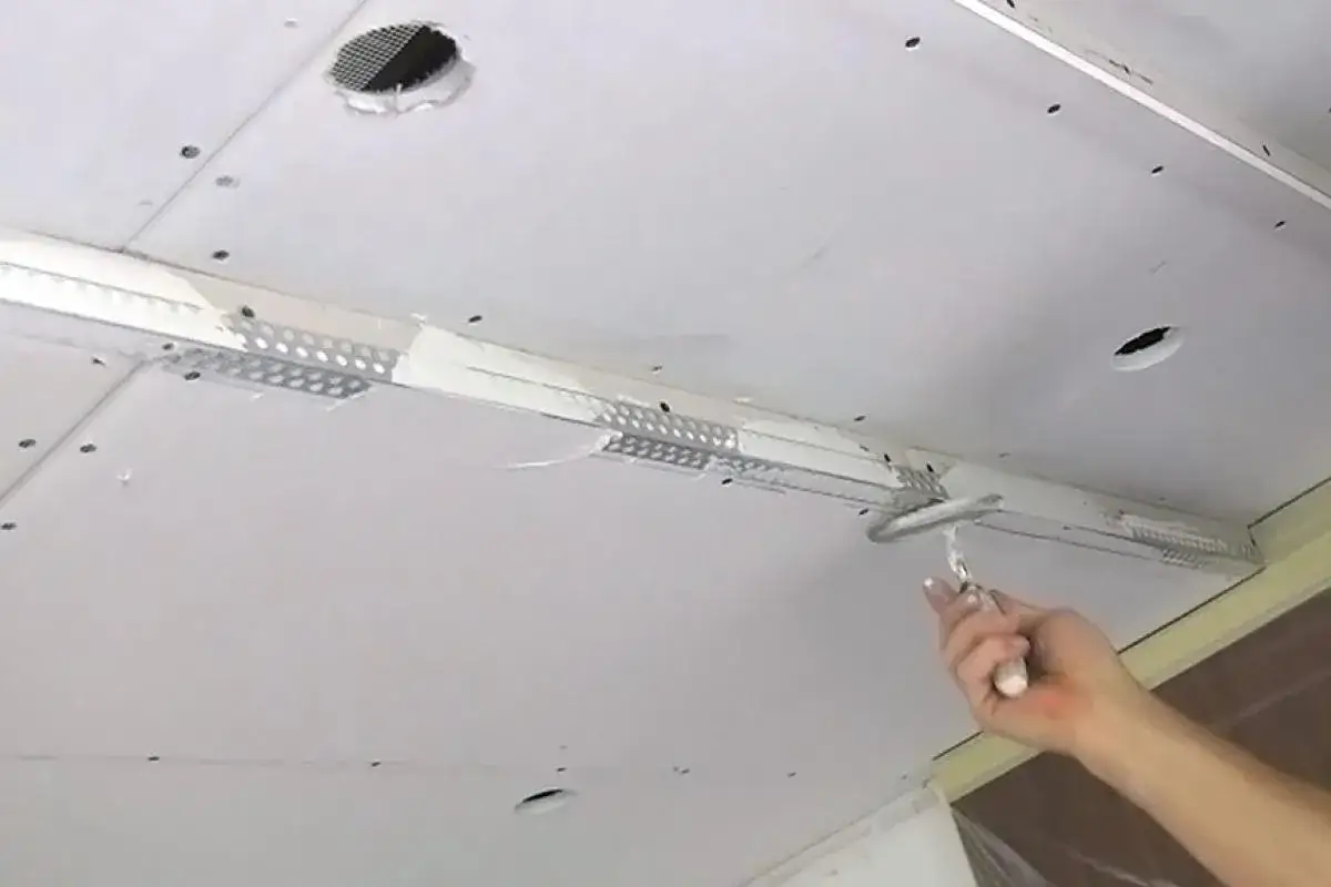 Hand verputzt eine Rigipsdecke mit einem Spachtel. Ein Lüftungsgitter ist sichtbar.