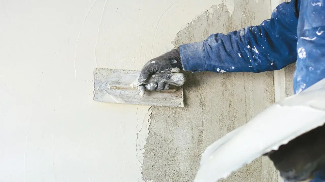 Hand mit Kelle tr&auml;gt Reibeputz auf eine Wand auf. Der Putz wird gleichm&auml;&szlig;ig verteilt, um eine glatte Oberfl&auml;che zu schaffen.