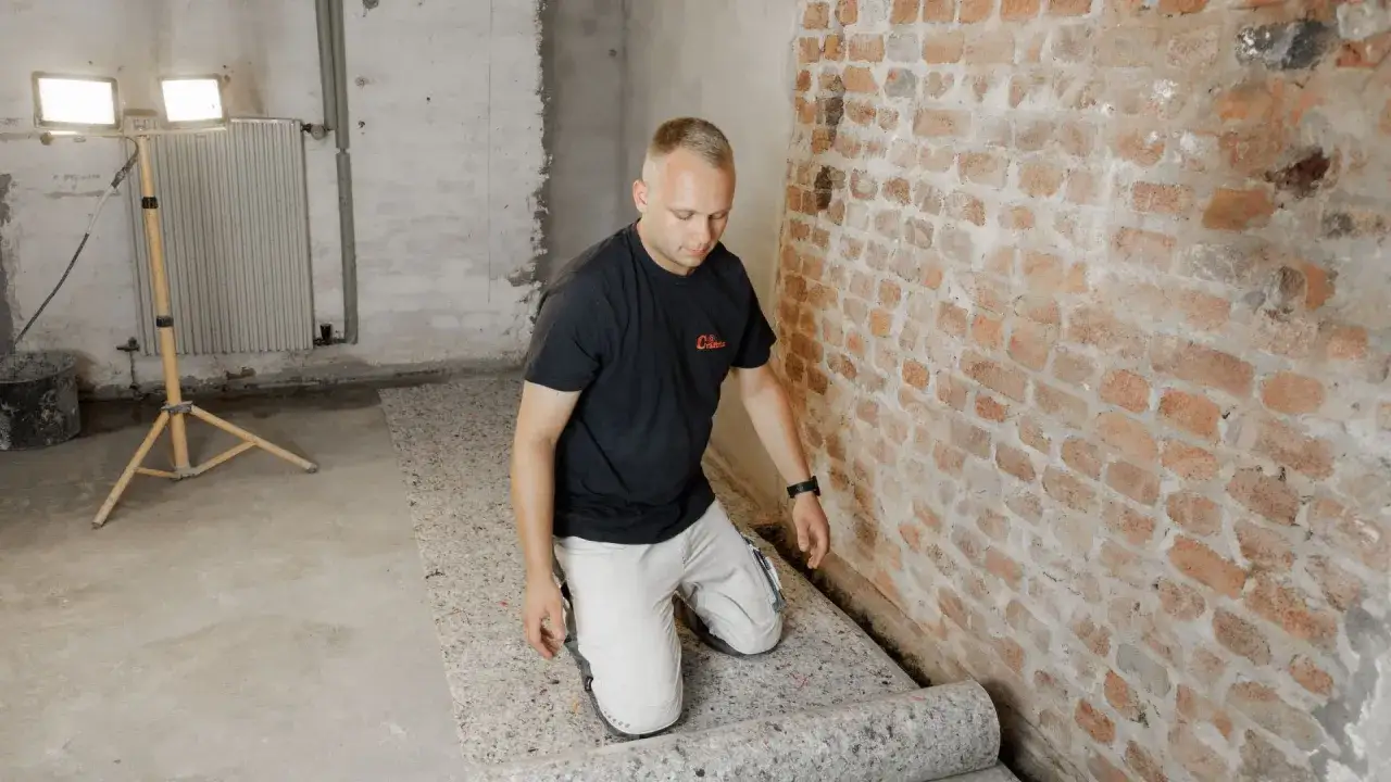 Mann kniet auf einem Bau-Boden und rollt eine D&auml;mmmatte aus. Er bereitet das Boden abdichten vor.