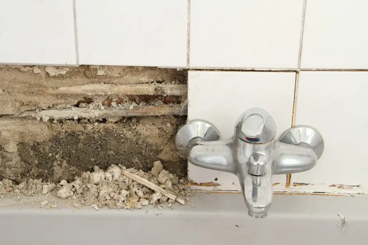 Ein Wasserrohr im Badezimmer ist freigelegt, was auf einen Wasserschaden hindeutet. Die Wandfliesen sind beschädigt, und Schutt liegt herum.